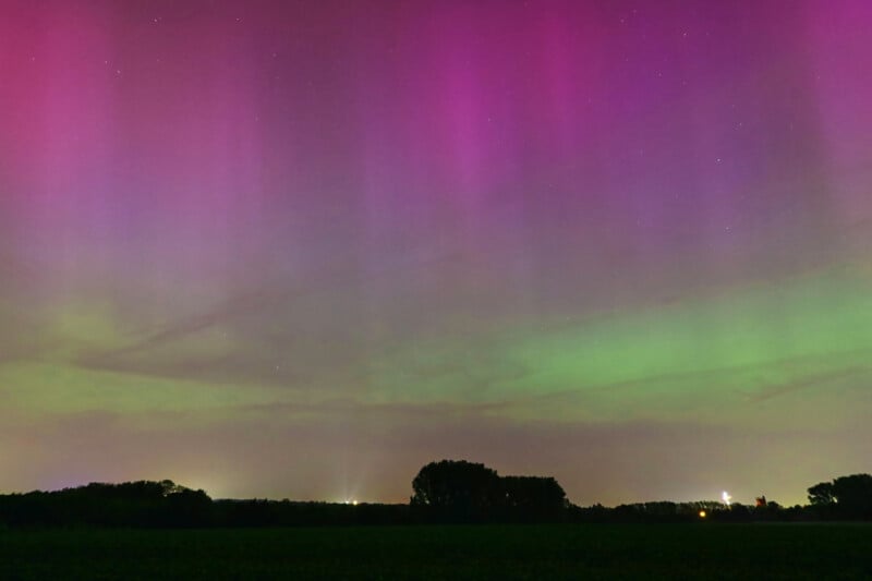 Night sky draped with the Northern Lights in shades of pink and green above a dark landscape with a silhouette of trees and faint lights on the horizon.