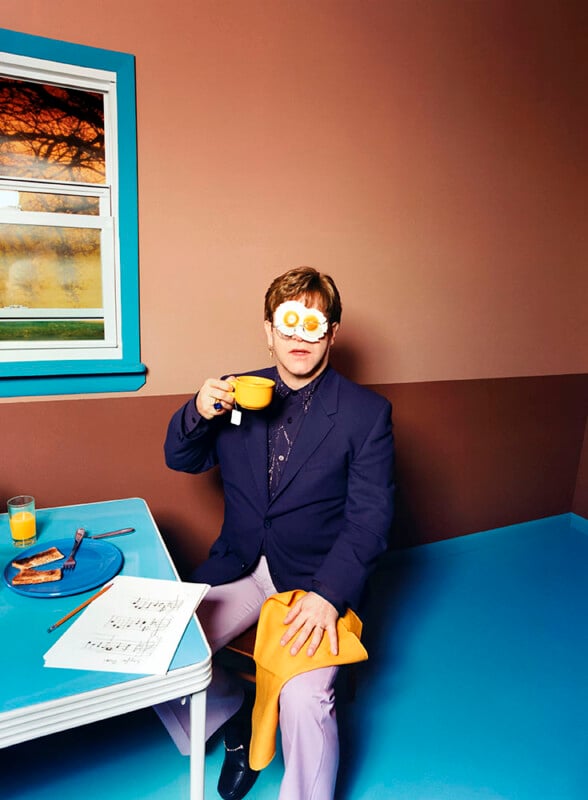 A person in a navy suit and light purple pants sits at a small table with a bowl of cereal in hand, wearing glasses shaped like fried eggs. The scene is in a room with brown walls and blue trim, with a table that has a newspaper, orange juice, and a plate of toast.