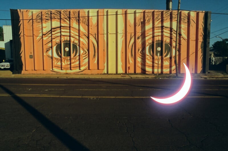 A large mural of two glaring eyes painted on an orange shipping container, with a neon pink crescent shape in front, set against a clear blue sky.