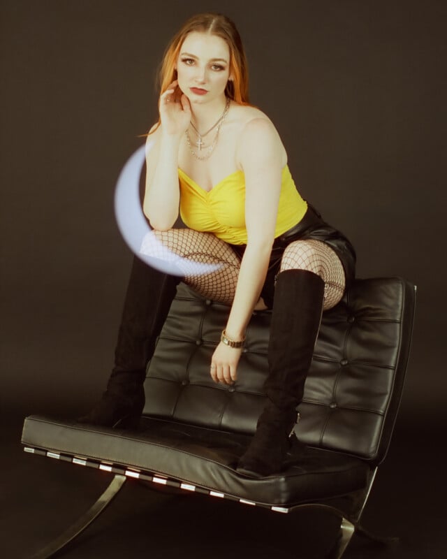 A woman with red hair sits thoughtfully on a black leather ottoman, wearing a yellow top, fishnet tights, and black boots, with a soft light orb effect near her face.