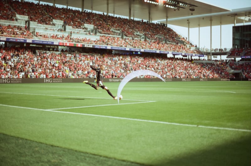 A soccer player strikes the ball during a corner kick in a packed stadium, showcasing a sunny day with a clear sky and the audience in vibrant red attire.