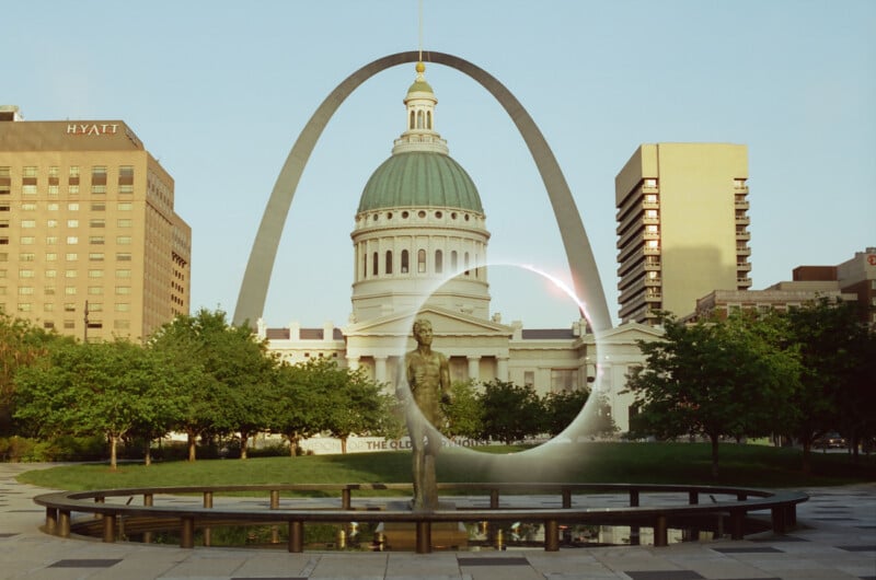 The gateway arch towers in the background with the old courthouse dome visible under it, framed by lush greenery and a bright blue sky. a statue stands prominently in the foreground.