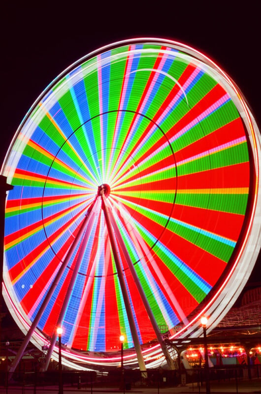 A ferris wheel captured in a long exposure at night, displaying a vibrant spectrum of swirling red, green, and blue lights against a dark sky.
