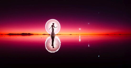 A silhouetted person in a flowing dress stands on a reflective, water-covered surface. Behind them, a glowing circle of light creates an ethereal effect. The sky is a gradient of deep purple to orange as the sun sets, with a few stars visible above.