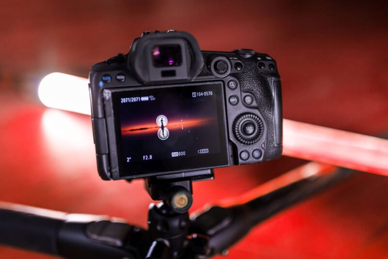A close-up of a digital camera on a tripod, focusing on a sunset scene displayed on its screen. A bright light source is visible behind the camera, creating a reflective glow on the red surface beneath.