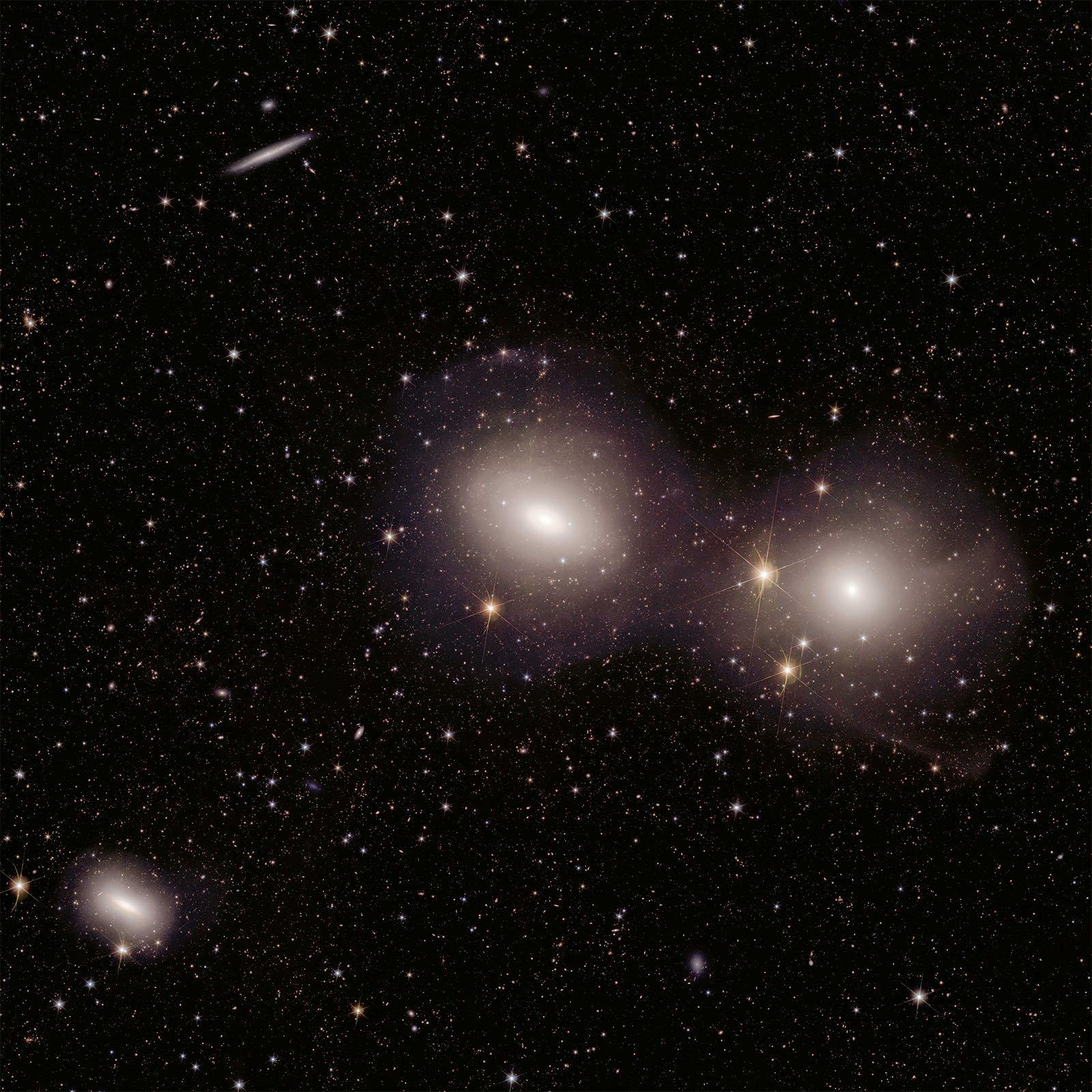 A deep space image showing two prominent, glowing galaxies in the center, surrounded by numerous stars and faint galaxies scattered across the dark expanse of the universe. The galaxies emit soft, diffuse light, contrasting against the vast, star-filled background.