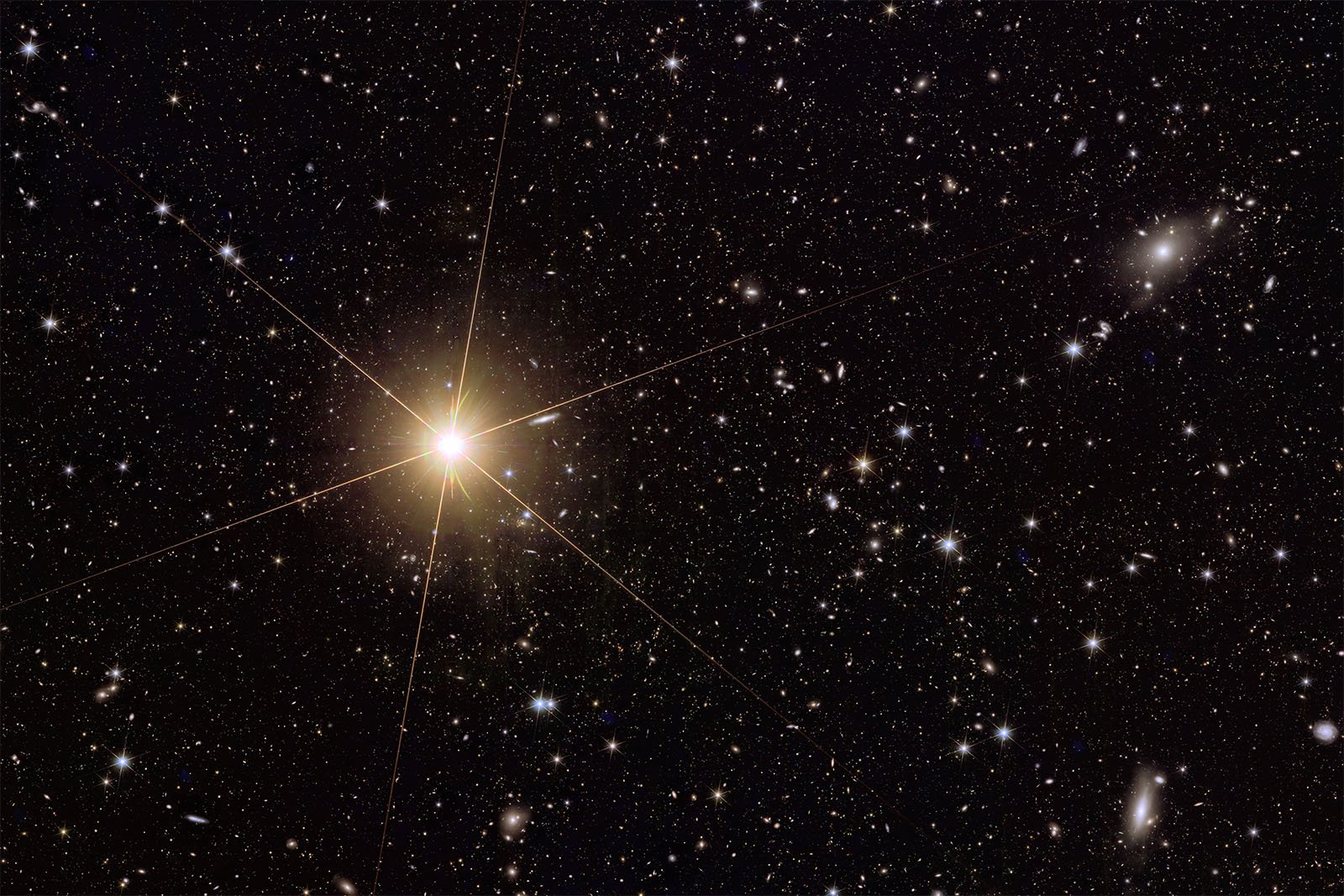 A bright yellow star shines in the center of a densely populated star field. The star emits prominent diffraction spikes, creating a cross-like pattern. Numerous other stars and galaxies of varying sizes and brightness are scattered across the dark backdrop of space.