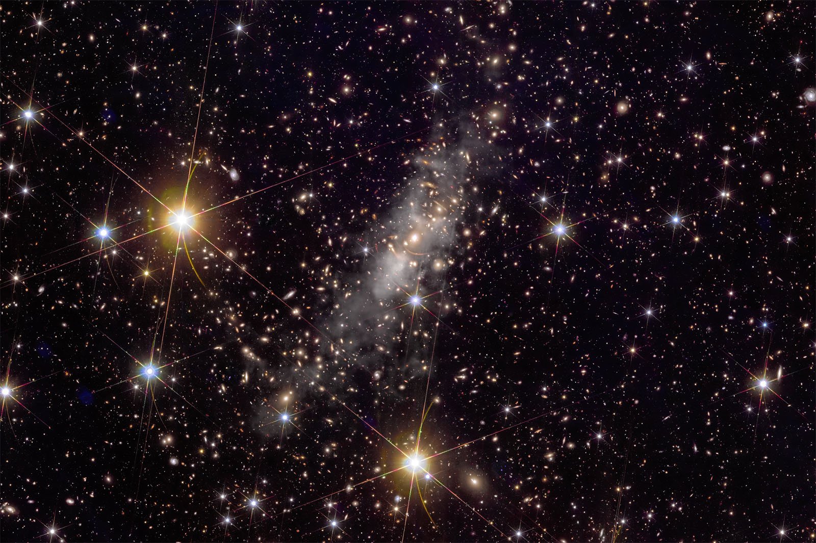 A dense field of stars fills the image, showcasing bright stars with diffraction spikes and many smaller stars scattered throughout. A hazy, elongated cloud of gas or dust stretches diagonally across the center, contrasting with the dark background of space.