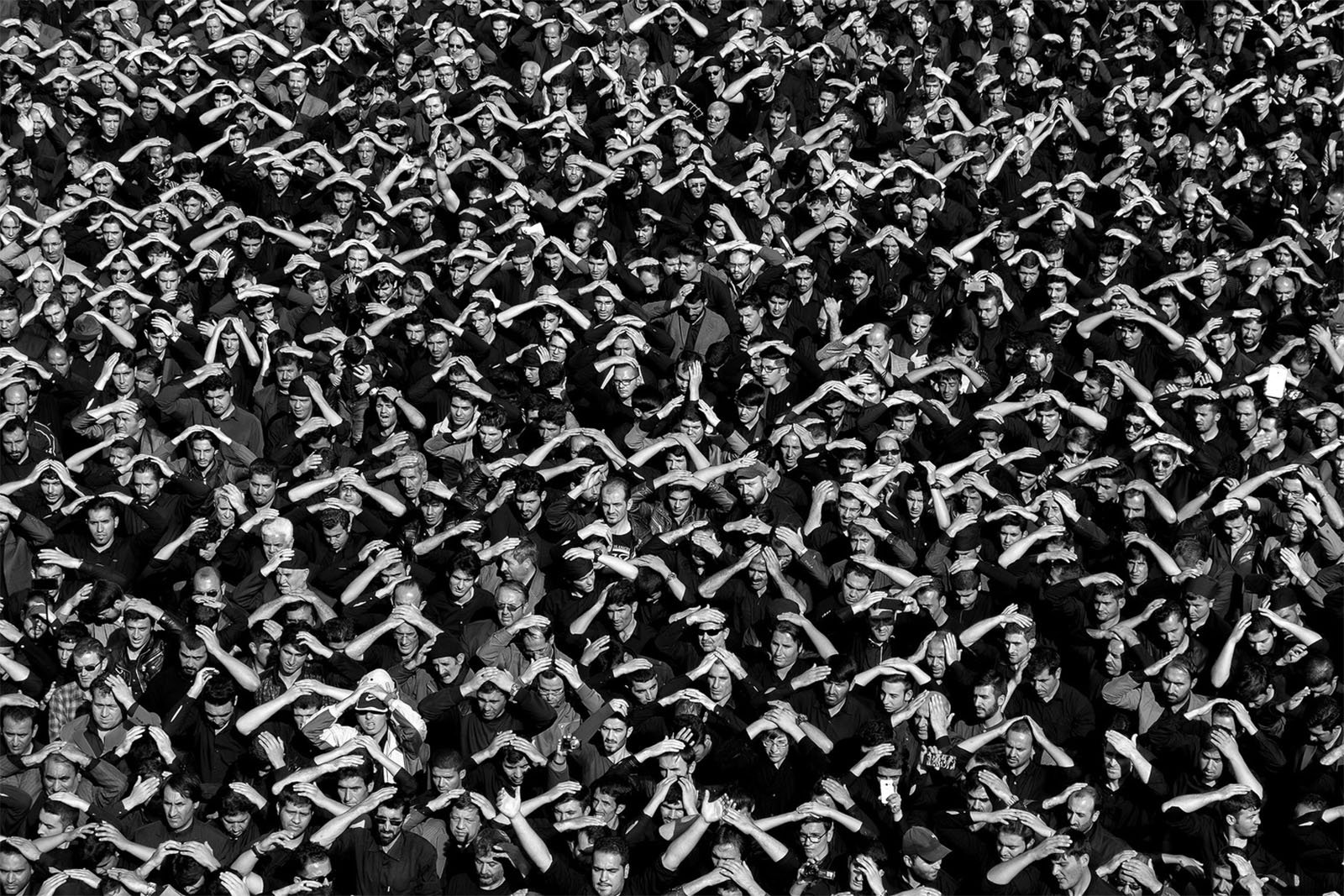 A large crowd of people, mostly dressed in dark clothing, standing close together with their hands raised and arms bent at the elbows in a uniform gesture. The image is in black and white, highlighting the dense and synchronized movement of the group.
