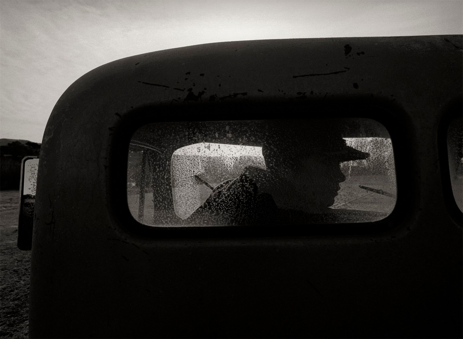 A silhouette of a person in a hat can be seen through a fogged and slightly dirty rear window of an old vehicle. The background is hazy, with barely discernible details, giving an atmospheric, mysterious feel to the black and white image.