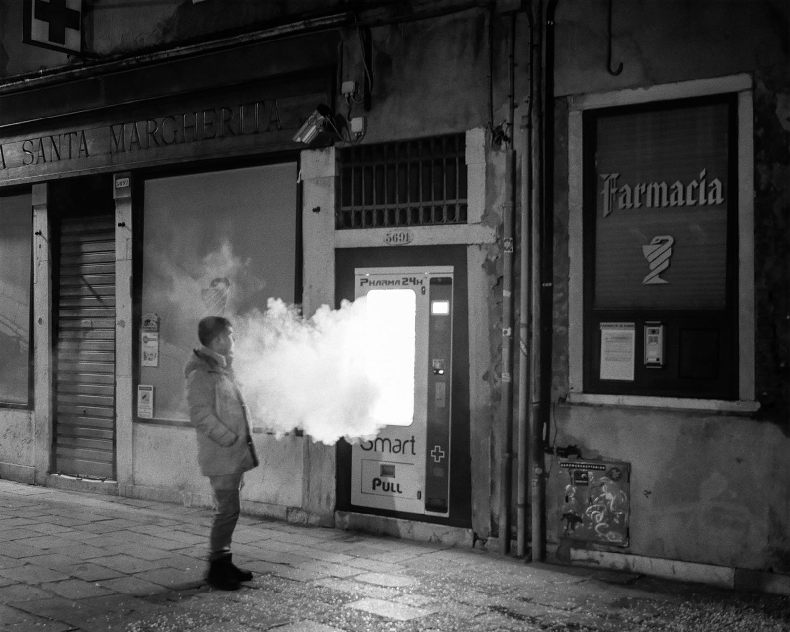 A person stands outside a closed pharmacy on a dimly lit street, exhaling a cloud of vapor. The scene is captured in black and white, emphasizing the contrast between the illuminated storefronts and the dark surroundings.