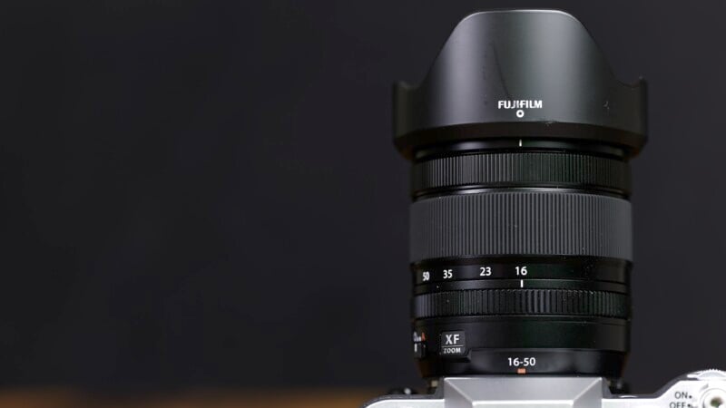 Close-up of a Fujifilm camera lens mounted on a camera body. The lens has a range of 16-50mm and features a lens hood attached. The background is dark, emphasizing the lens and its markings.