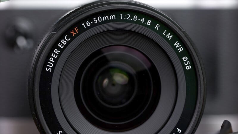 Close-up image of a camera lens displaying the text "SUPER EBC XF 16-50mm 1:2.8-4.8 R LM WR Ø58" around the rim. The lens is prominently centered and captures intricate details, with a blurred background hinting at the camera body.