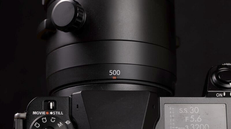 Close-up image of a black camera lens with a focus ring marked "500." The top part of the camera shows a knob and part of the body, while the lower part displays an LCD with various settings, showcasing its professional and detailed design.