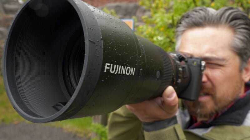 A person is holding and aiming a Fujinon camera with a large telephoto lens, partially obscuring their face. The camera lens is slightly wet, indicating a recent rain. The background features green foliage and hints of stone structures.