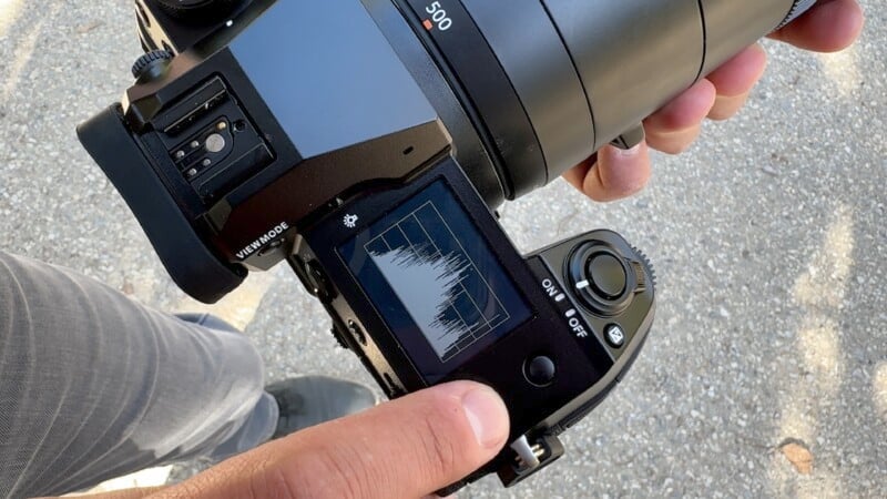 A person holding a digital camera with a flip screen displaying a histogram and various settings, viewed from above. The background shows a paved surface.