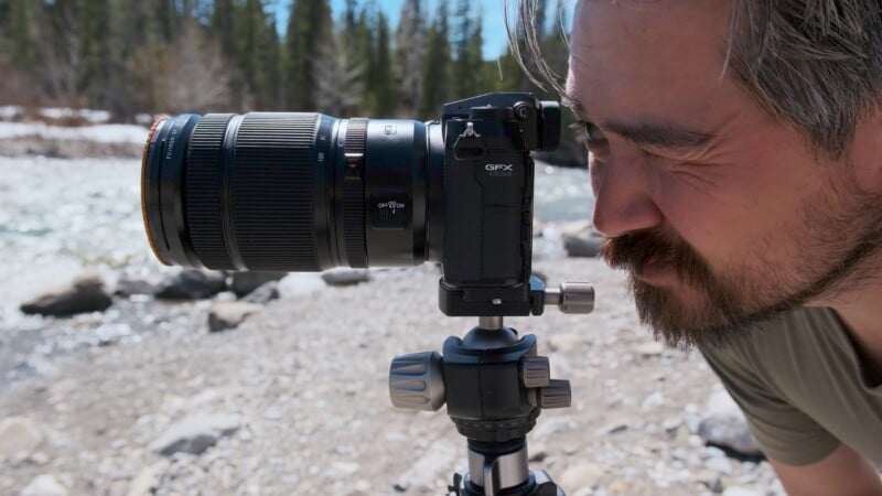 A man with a beard closely examining the viewfinder of a large DSLR camera mounted on a tripod, set against a background of a sunny, rocky riverbank.