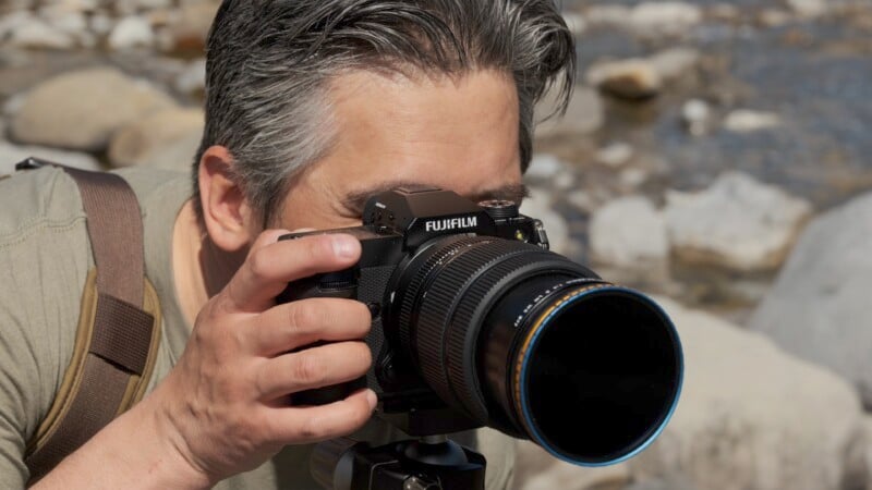 A man with gray hair using a Fujifilm camera to take photos outdoors, focusing intently on his subject. He's wearing a beige backpack and is surrounded by rocks and a riverbed.