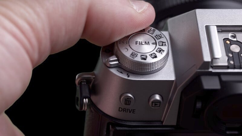 A close-up image showing a person's thumb adjusting the mode dial on a professional camera from 'Drive' to 'Film'.