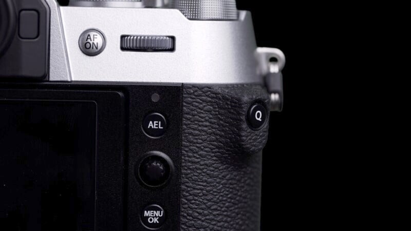 Close-up of a modern digital camera focusing on its controls, including the AF ON, AE-L, and menu buttons, with a textured grip, against a dark background.