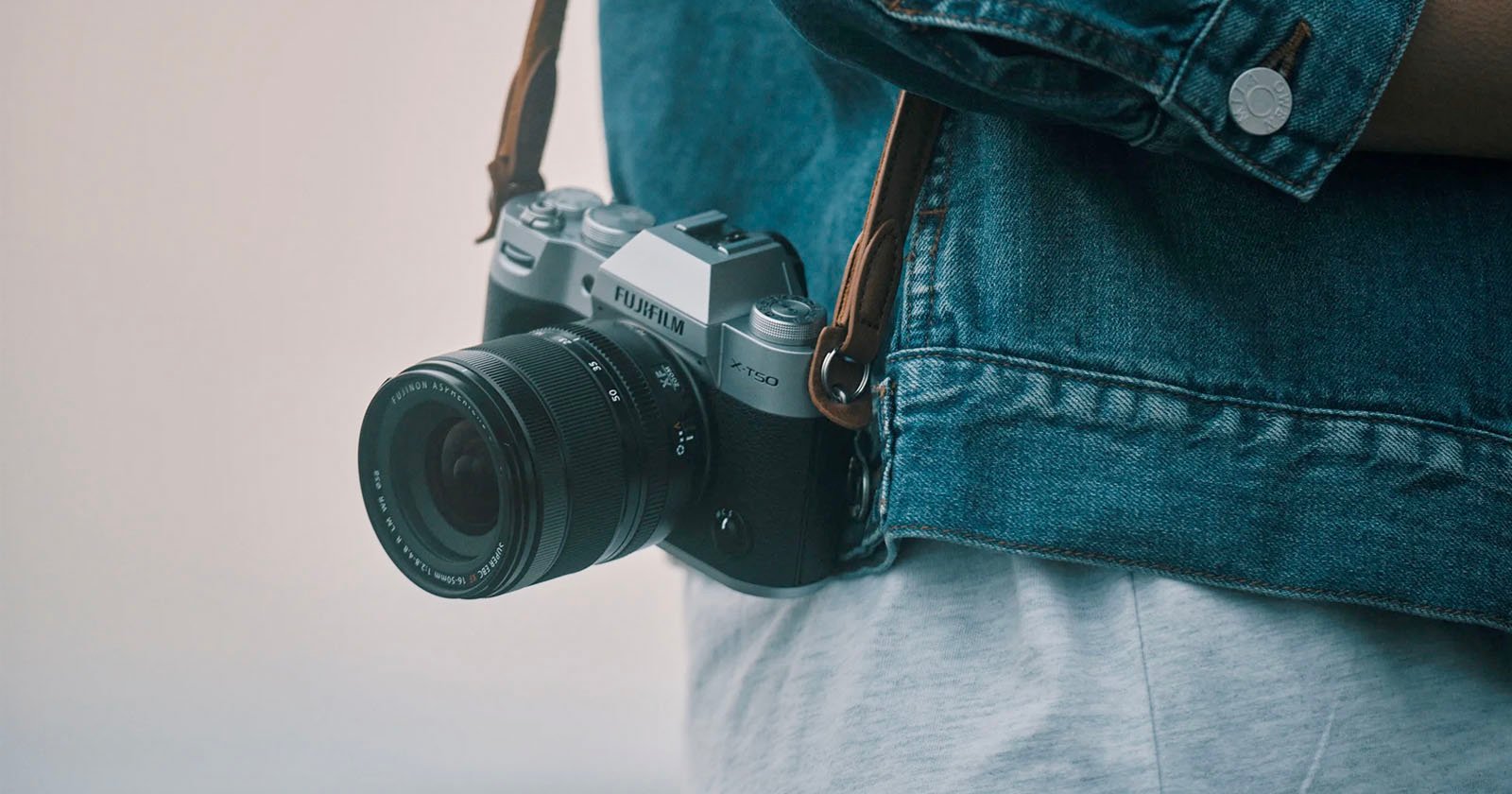 A person wearing a denim jacket has a Fujifilm XT20 camera hanging around their neck. The camera is silver and black with a lens attached, and it is secured by a brown strap. The background is blurred, focusing attention on the camera and the jacket.