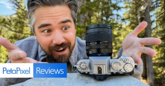 A person with a beard and mustache enthusiastically gestures towards a DSLR camera set on a rock. The background features a forest scene. There is a "PetaPixel Reviews" logo in the bottom left corner of the image.