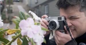 A man focuses intently while taking a close-up photograph of pink flowers using a vintage-style camera, with blurred buildings in the background.