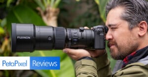 A person with gray hair and a beard is looking through the viewfinder of a camera with a large Fujinon lens. The setting appears to be outdoors with green foliage in the background. The image includes the text "PetaPixel Reviews" in the bottom left corner.