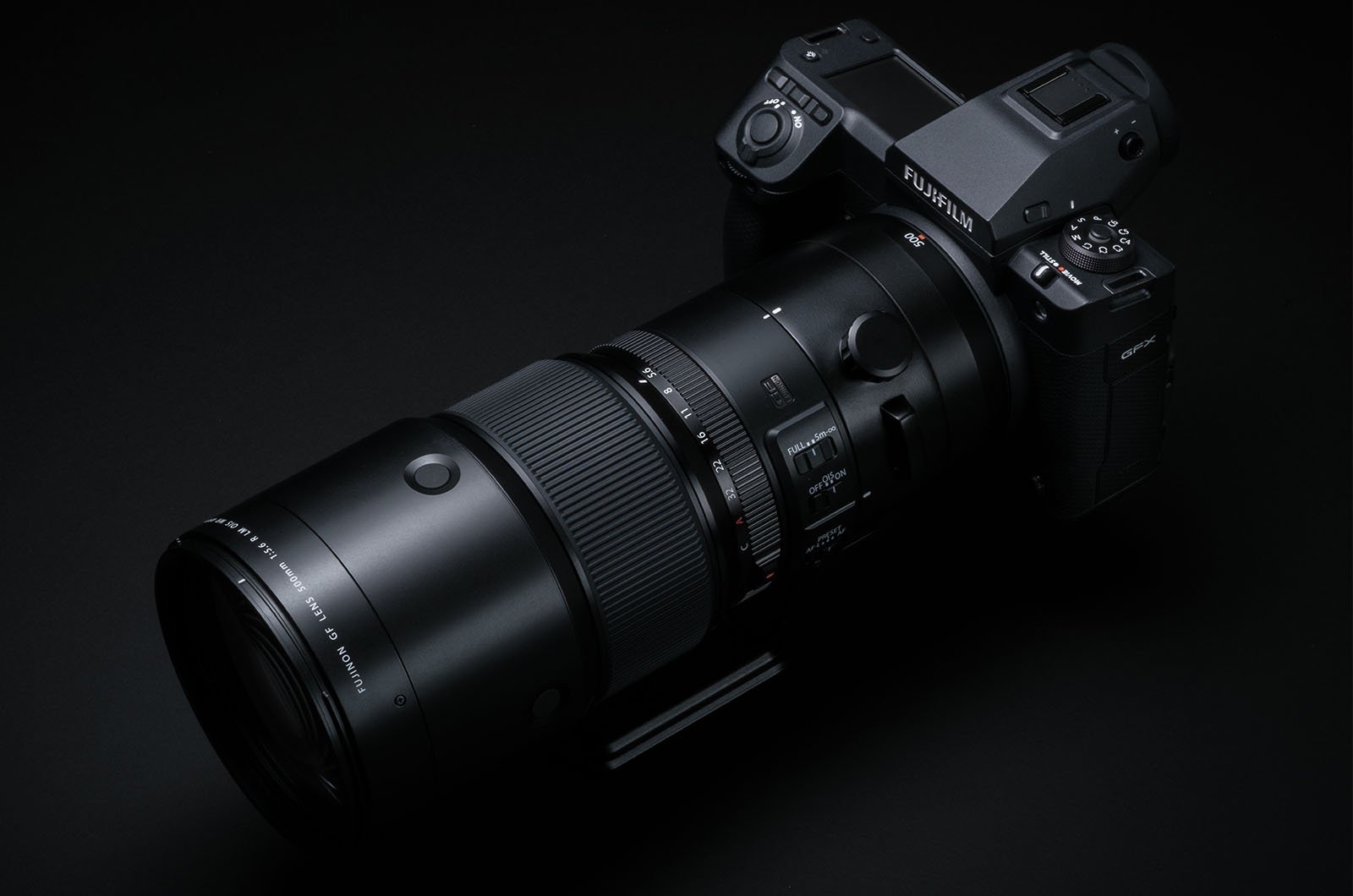 A sleek black Fujifilm camera with a large, extended zoom lens attached is displayed on a black surface with a black background. The camera features various control dials, buttons, and a digital screen, highlighting its professional-grade design.