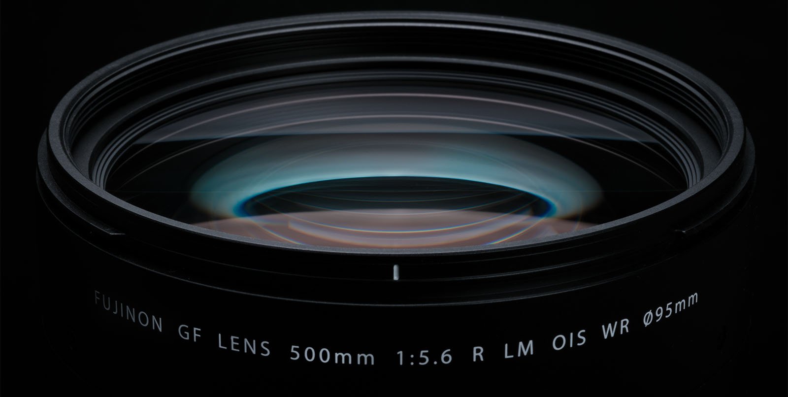 Close-up view of a camera lens showing the glass elements and reflecting light. The text on the lens reads "FUJINON GF LENS 500mm 1:5.6 R LM OIS WR Ø95mm." The image highlights the intricate details of the lens construction.
