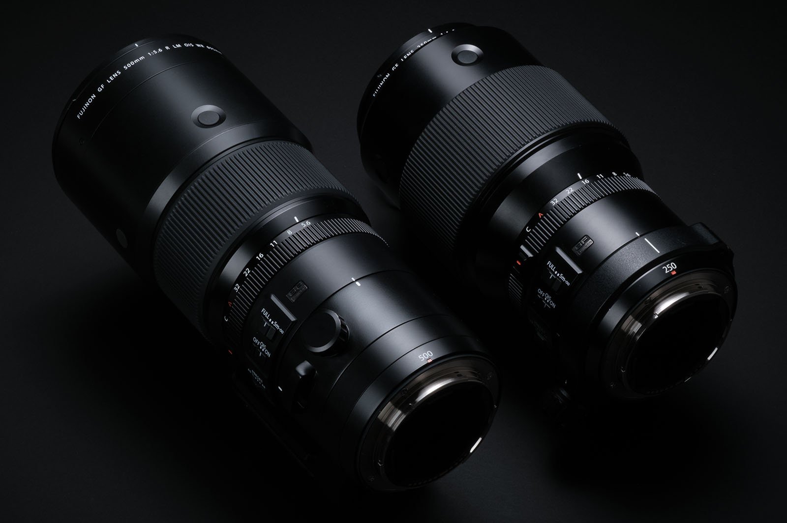 Two black, professional camera lenses are placed side by side on a dark background. The lenses are sleek and feature various control dials and focus rings. The lens on the left is slightly larger than the one on the right.