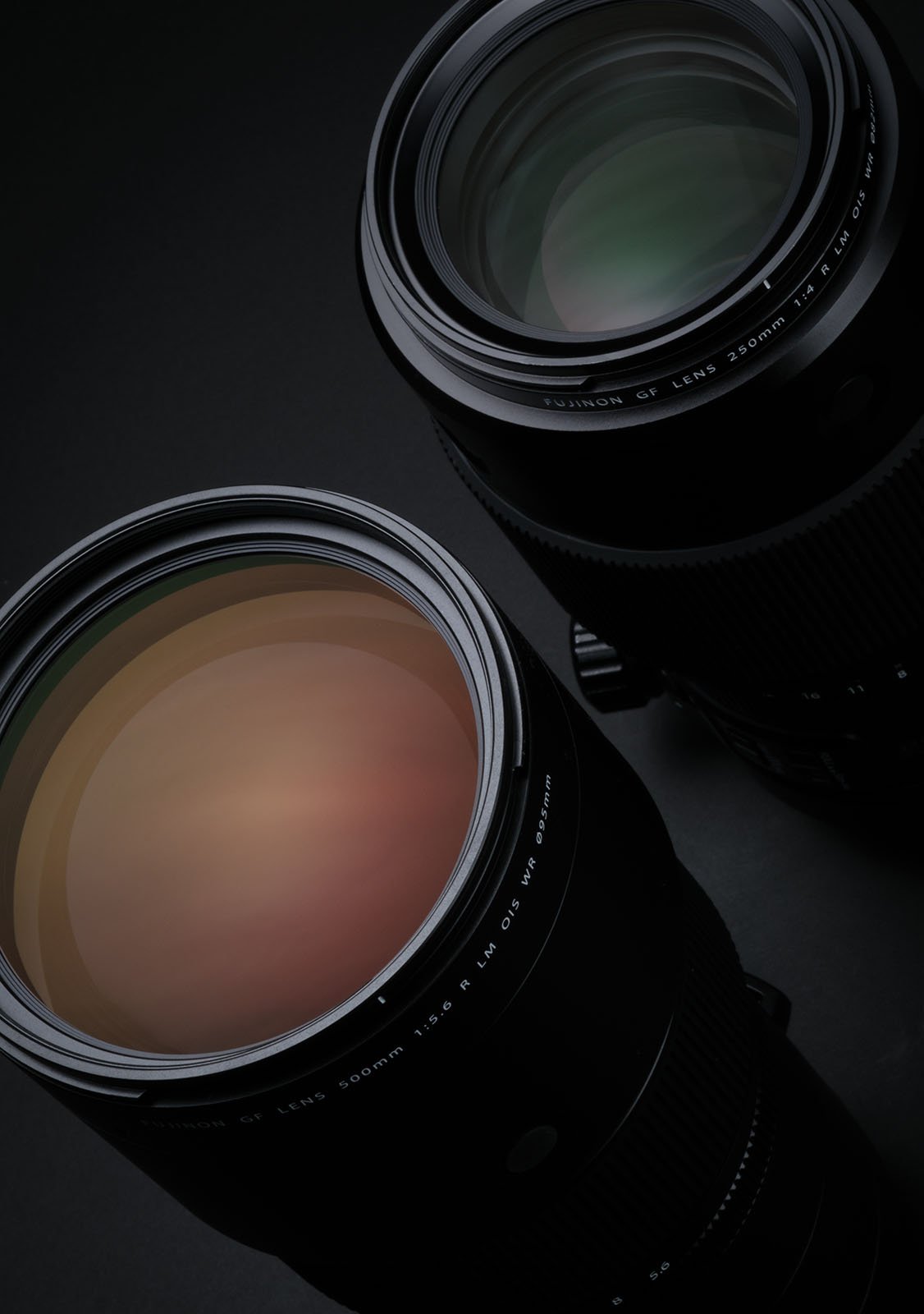 Close-up view of two camera lenses with large glass elements, positioned diagonally against a dark background. The lenses, reflecting subtle hues of green and orange, showcase their intricate design details and barrel markings.