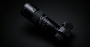 A black DSLR camera with an attached long zoom lens is placed on a black surface, partially illuminated to highlight its details. The camera and lens have a sleek and professional design, creating a striking contrast against the dark background.