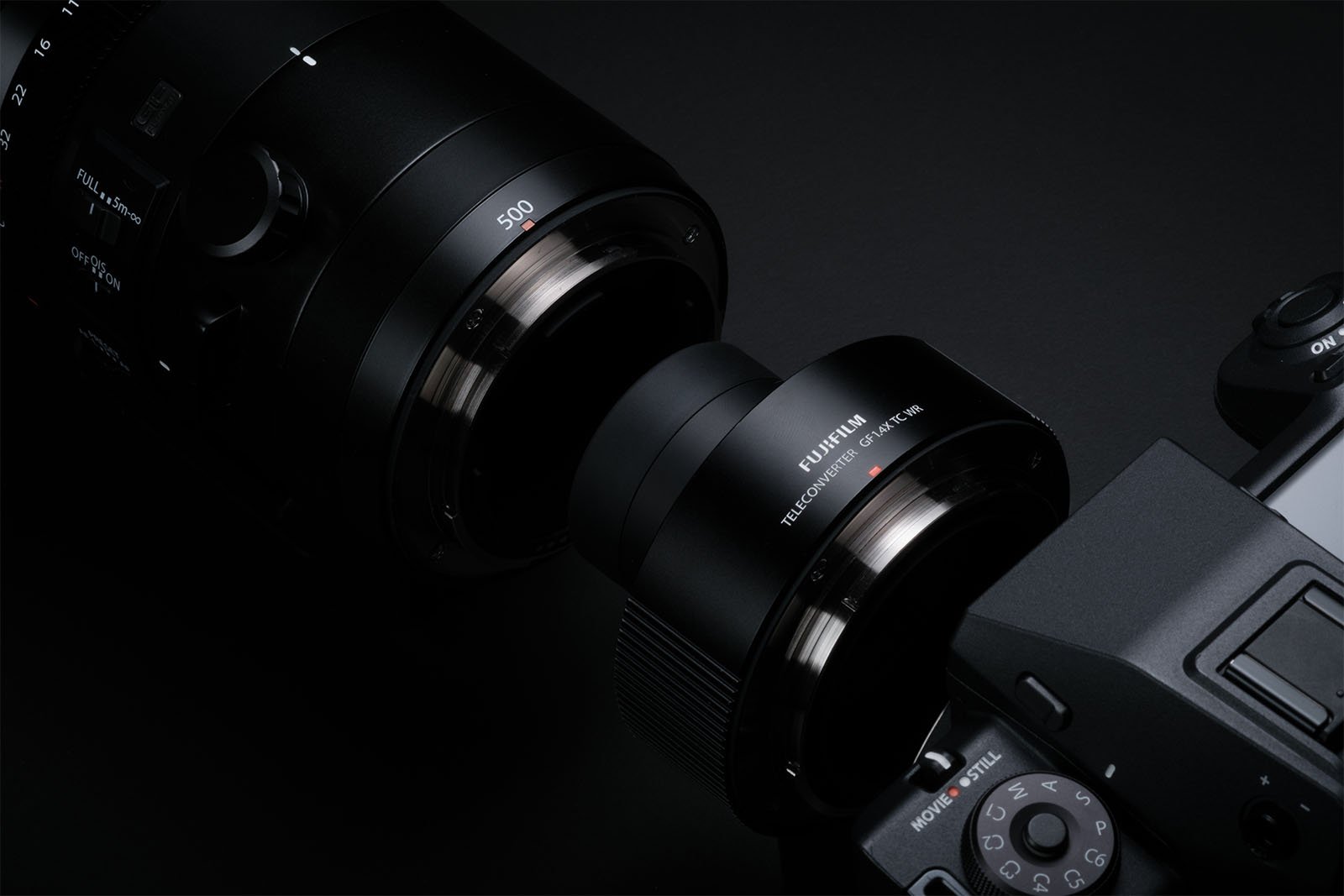 A close-up shot of a Fujifilm macro extension tube connected between a camera body and a lens on a black background. The metallic connections and labels on the tube and lens are visible, showcasing the brand and model details.