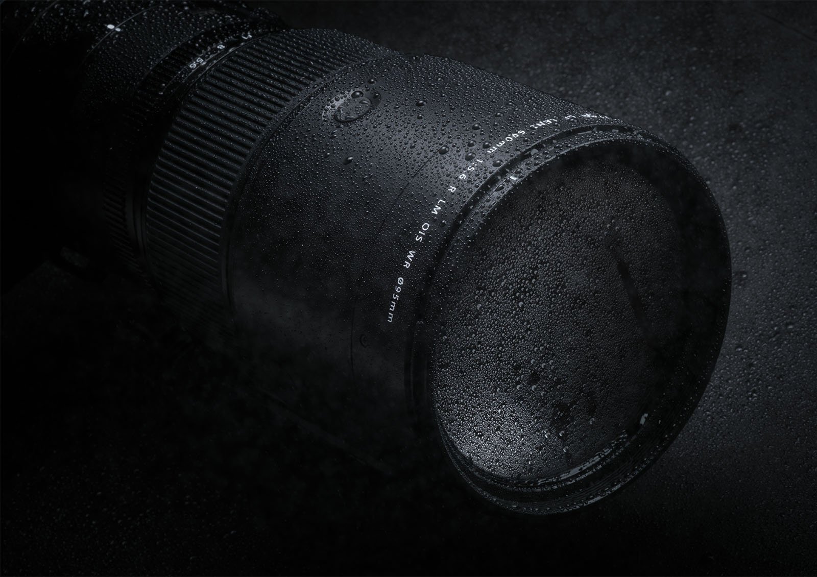 A close-up shot of a camera lens covered in water droplets. The lens, shown in a dark and moody setting, highlights its sleek and rugged design, suggesting it is weather-sealed and durable enough to withstand harsh conditions.