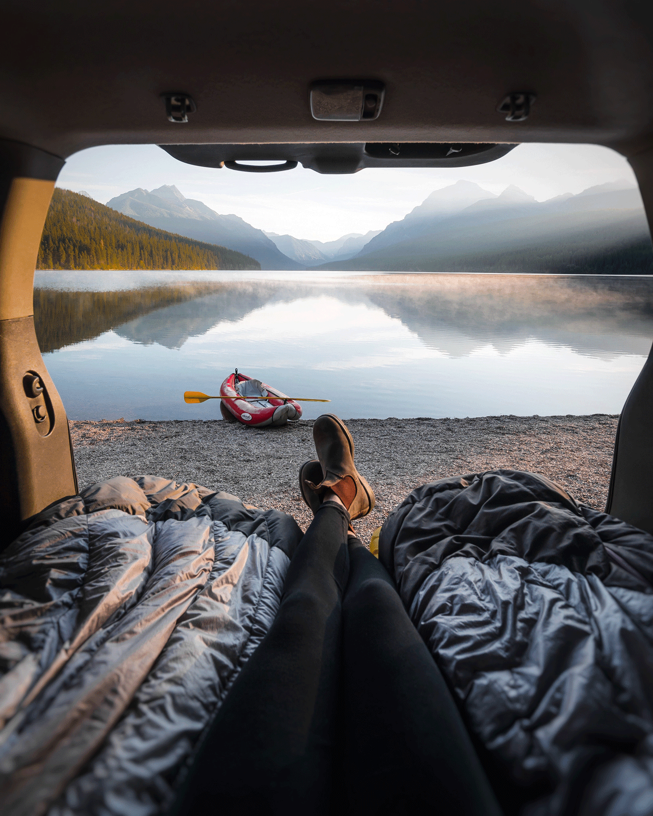 A person is lying in the back of a car with the trunk open, looking out at a serene lake surrounded by mountains. A kayak with a paddle is on the shore near the water. It's a peaceful outdoor scene with mist over the lake.