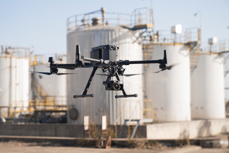A black quadcopter drone equipped with a camera hovers in front of industrial storage tanks with metallic pipes and railings. The sky is clear and the background features large white cylindrical containers, likely part of an industrial facility.