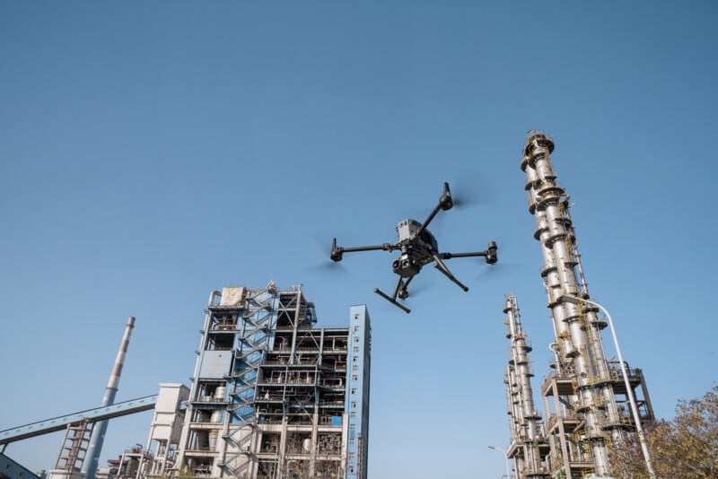 A drone hovers in the sky near an industrial facility with tall metal structures and large machinery against a clear blue sky. The drone is equipped with a camera and is flying in close proximity to the towers and pipes of the plant.
