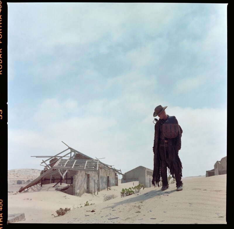 A person dressed in dark, tattered clothing, with a hat and backpack, stands in a desert-like area near a dilapidated wooden structure. The sky is overcast, and the scene has a post-apocalyptic feel.