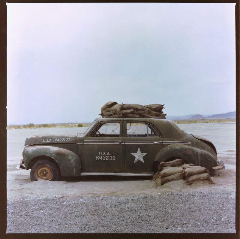 A vintage military car with "U.S.A 19422122" marked on its side is stuck in mud. Sandbags are piled on the roof and around the vehicle. The car has a white star painted on the side door. The background features a desolate, flat landscape under an overcast sky.