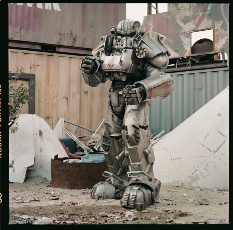 A person is wearing an imposing, metallic power armor suit, standing in a scrapyard surrounded by scattered debris, rusty barrels, and large wooden and metal structures. The suit features large shoulder pads, intricate mechanisms, and a helmet with a visor.