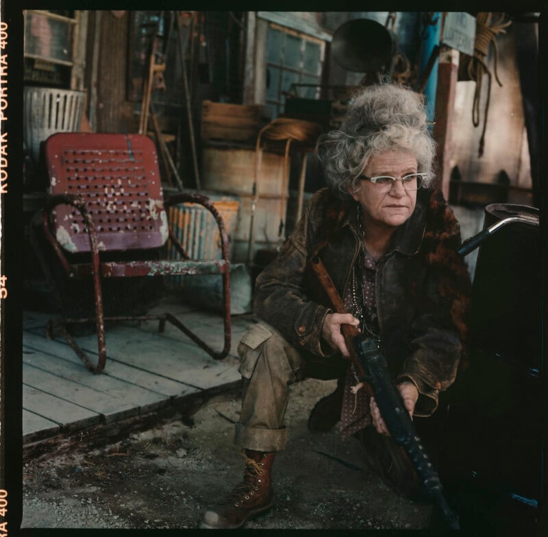 An elderly woman with gray, curly hair, glasses, and rugged clothing holds a rifle while kneeling outside a dilapidated building. She appears alert and cautious. The scene includes an old, rusty metal chair and various worn objects in the background.