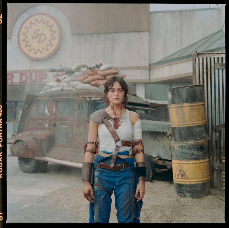 A person stands outside a run-down building with a damaged, armored vehicle in the background. They are wearing a rugged, post-apocalyptic outfit with arm and shoulder armor and utility belts. Their clothing and skin are smeared with what appears to be blood.