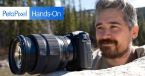 A close-up shot of a Fujifilm GFX100S camera with a large lens resting on a rock, with a blurred background of trees and water. The PetaPixel logo and the words "Hands-On" are displayed in the top left corner. A person with a beard is looking at the camera.