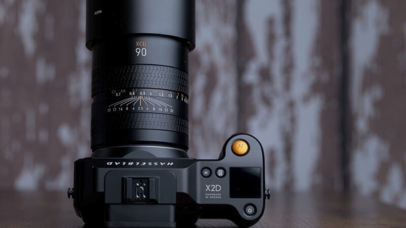 A hasselblad x1d camera with a 90mm lens attached, on a wooden surface with a blurry background showing wall textures. the focus is on the lens markings and camera controls.