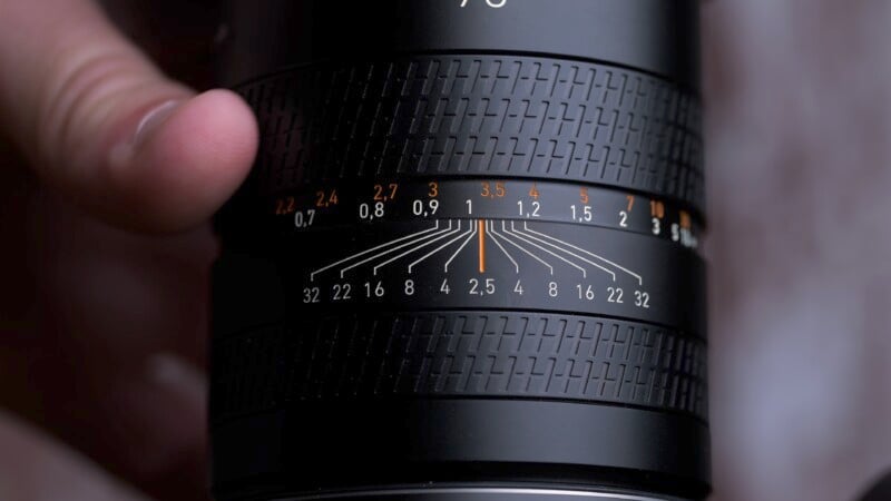 A close-up photo of a camera lens held by a hand, focusing on the aperture and focus scale marked with numbers and lines, highlighting the depth of field.