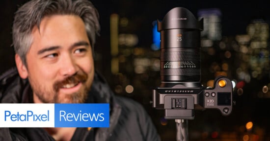 A man with a slight smile looks at a camera mounted on a tripod in the foreground, with a city's blurry lights in the background. the image includes a "petapixel reviews" label.