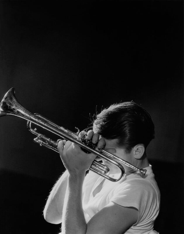 A black-and-white photo shows a person playing a trumpet. The individual is holding the trumpet high, with the mouthpiece pressed to their lips. They are wearing a light-colored short-sleeve shirt, and their face is partially obscured by their hands and the instrument.
