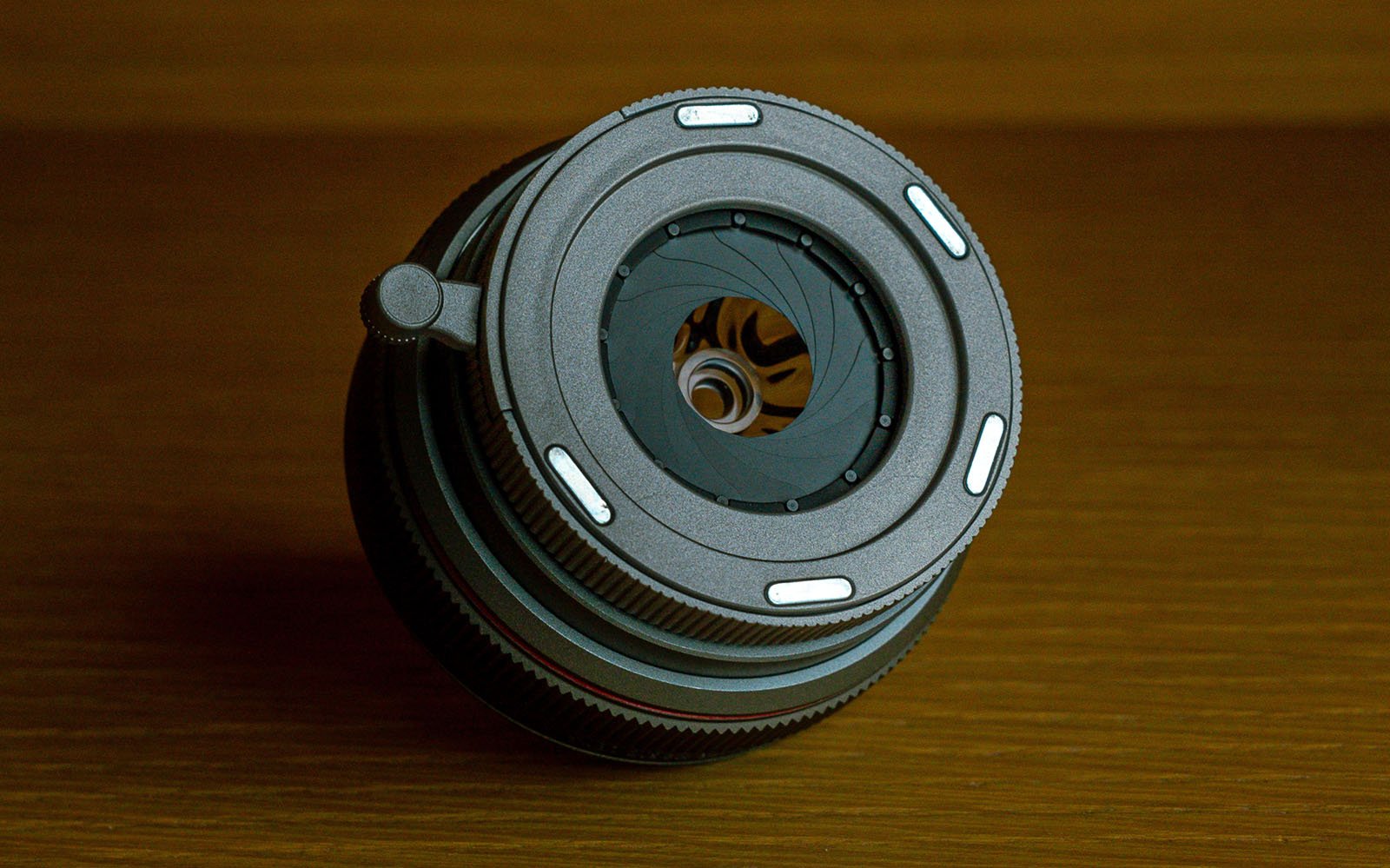 A close-up image of a camera lens placed on a wooden surface. The lens is positioned facing upwards, showcasing the aperture blades and intricate inner components. The background appears warm and softly lit.