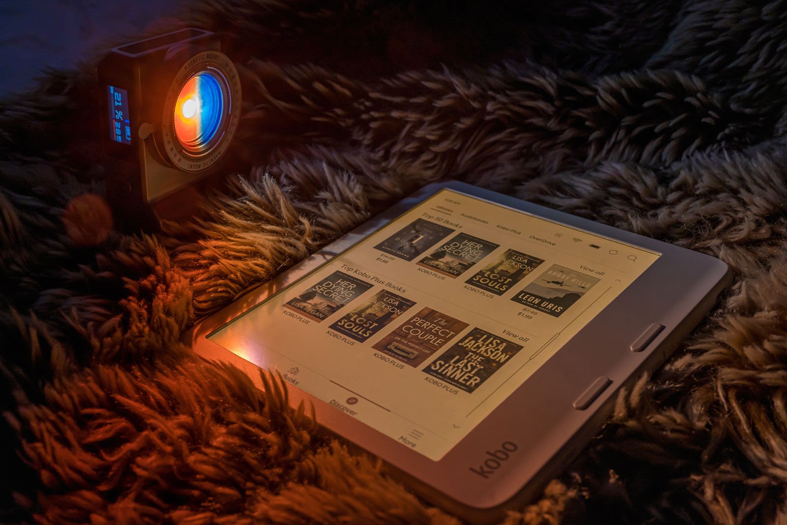 A Kobo e-reader displays a selection of books while resting on a fluffy, dark-colored surface. A vintage-style camera with a glowing blue light lens is positioned nearby, casting an orange glow and creating a warm, cozy atmosphere.